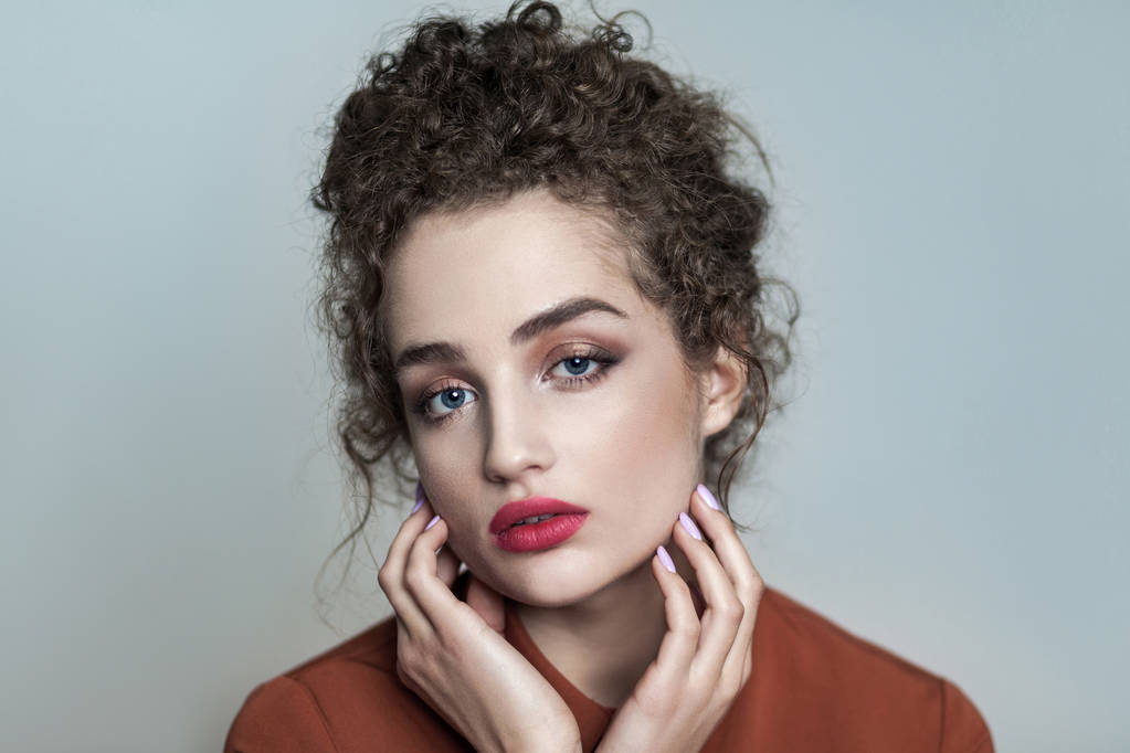 Schoonheid portret van jonge mooie model met verzamelde donker krullend haar en naakt make-up met rode lippen in bruin shirt camera kijken en ontroerend gezicht op grijze achtergrond  - Foto, afbeelding