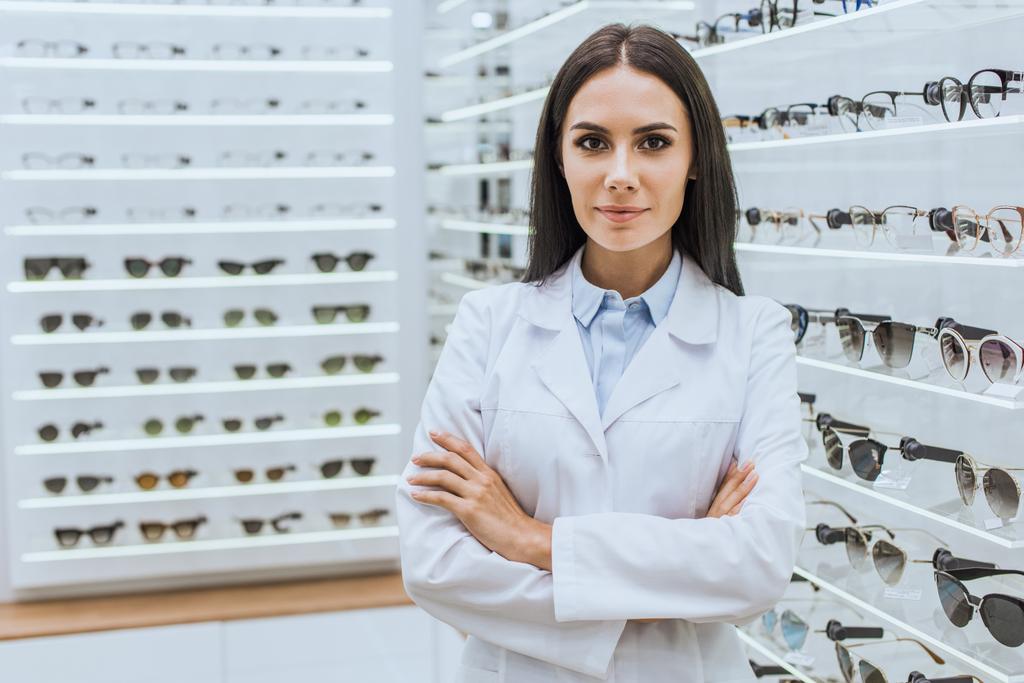 optometrista profesional con brazos cruzados posando cerca de estantes con vista en óptica
 - Foto, Imagen