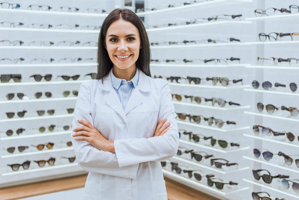 profissional optometrista sorridente posando com braços cruzados perto de prateleiras com visão na óptica
 - Foto, Imagem