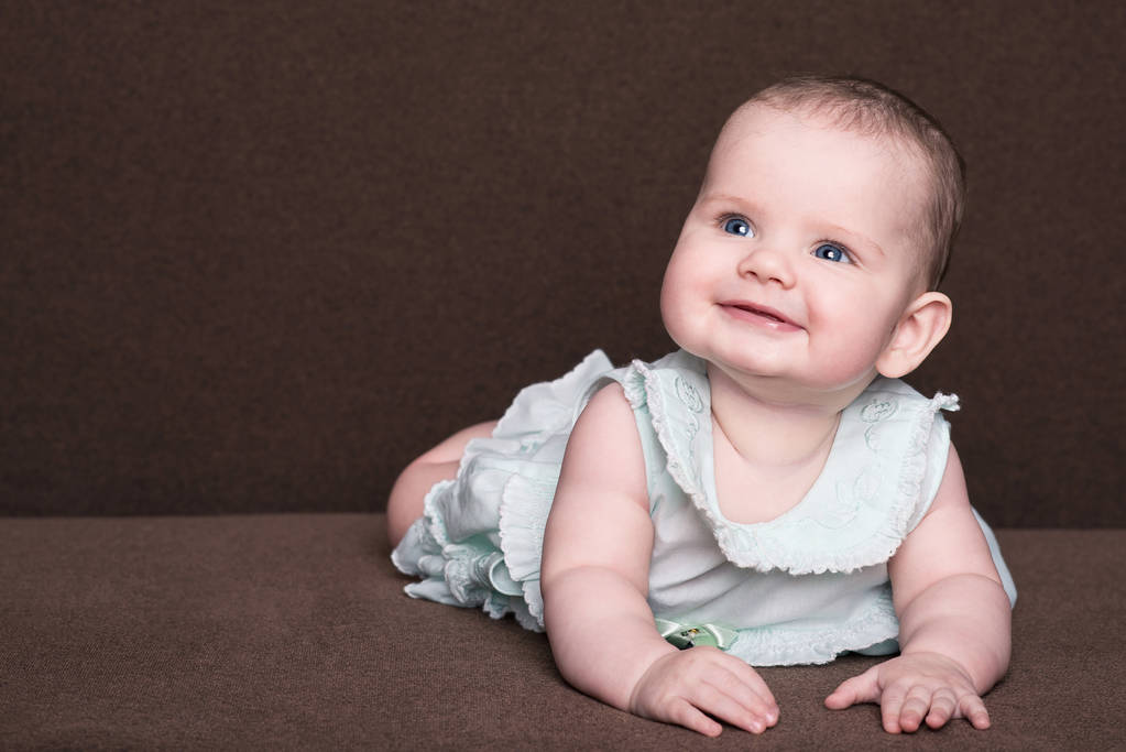 sonriente niña en vestido de verano con grandes ojos azules se encuentra en su vientre sobre fondo marrón
 - Foto, Imagen