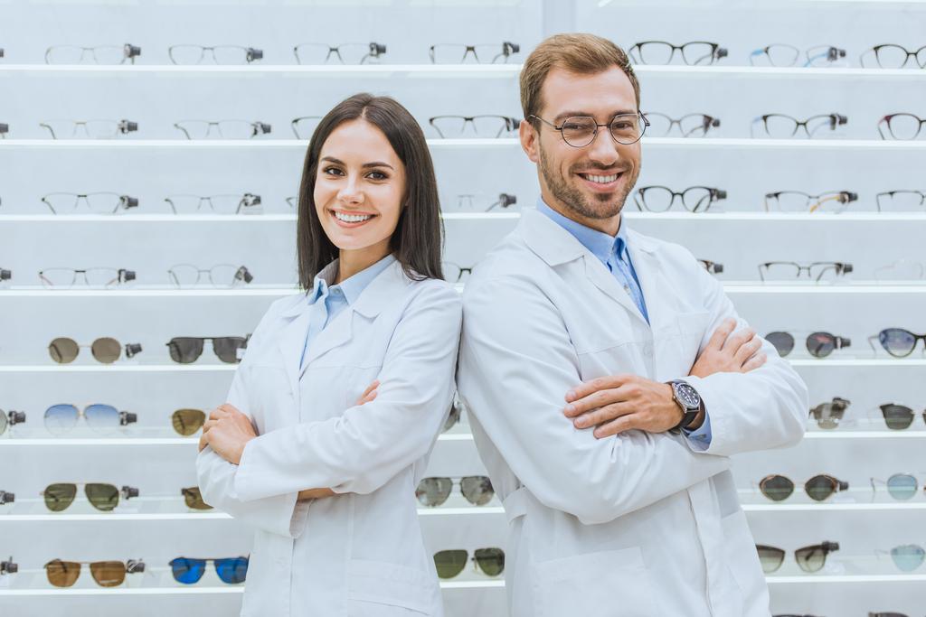 профессиональные оптики, позирующие со скрещенными руками в офтальмологическом магазине с очками
 - Фото, изображение