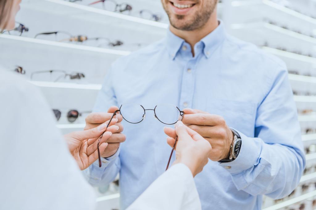 cropped image of female oculist giving eyeglasses to young man in ophthalmic shop - Photo, Image
