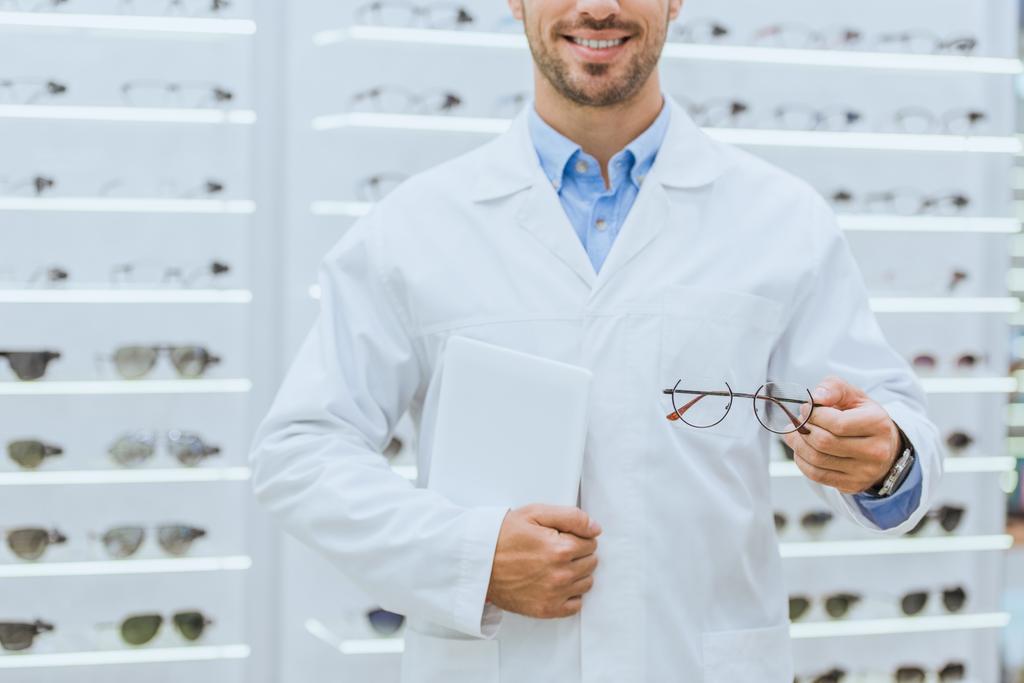 Ausgeschnittene Ansicht des Augenoptikers in weißem Kittel mit Brille und digitalem Tablet in Optik - Foto, Bild