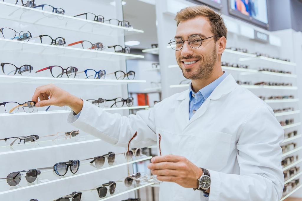 Lächelnder Optiker im weißen Kittel nimmt Augenlicht aus Regalen in der Augenheilkunde - Foto, Bild