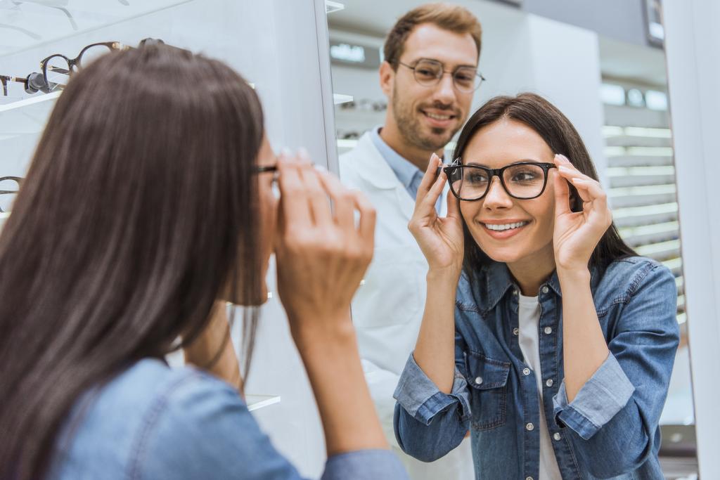 Rückansicht einer fröhlichen Frau, die eine Brille wählt und in den Spiegel schaut, während der männliche Augenarzt in der Nähe in der Optik steht  - Foto, Bild