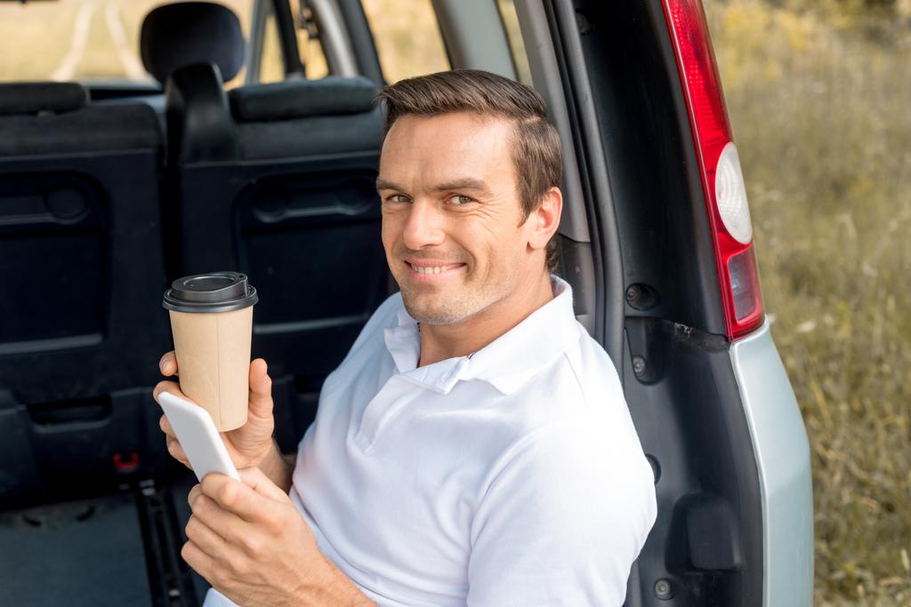車のトランクに座っていると、スマート フォンを使用しているときのカメラ目線のコーヒーの紙コップを持つ幸せな男 - 写真・画像