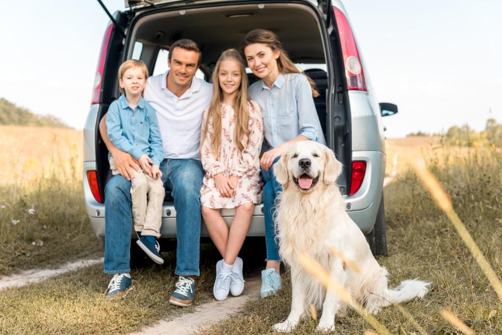 ευτυχισμένος νεαρή οικογένεια με retriever σκύλου συνεδρίαση στο πορτ-μπαγκάζ αυτοκινήτου και βλέπουν φωτογραφική μηχανή στο πεδίο - Φωτογραφία, εικόνα