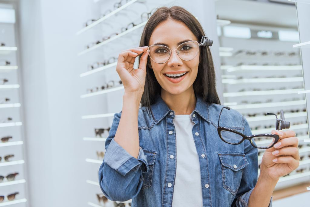 εύθυμη νεαρή γυναίκα επιλέγοντας γυαλιά και βλέπουν φωτογραφική μηχανή σε οφθαλμικά κατάστημα - Φωτογραφία, εικόνα