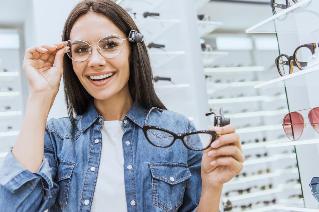 портрет привлекательной счастливой женщины, выбирающей очки и смотрящей в камеру в офтальмологическом магазине
 - Фото, изображение