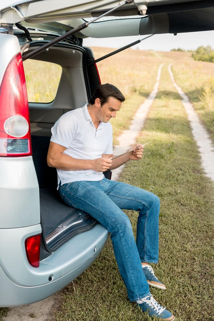 handsome smiling man sitting in car trunk and looking at map in field - Photo, Image