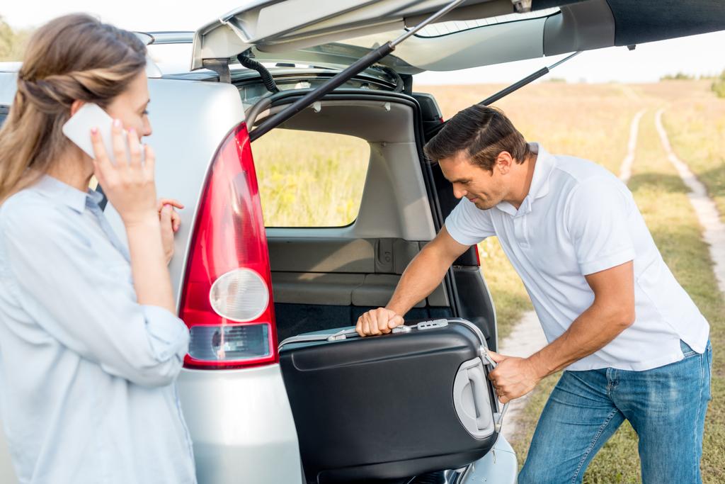 heureux adulte l'emballage des bagages dans le coffre de la voiture tandis que sa petite amie parle par téléphone dans le domaine
 - Photo, image