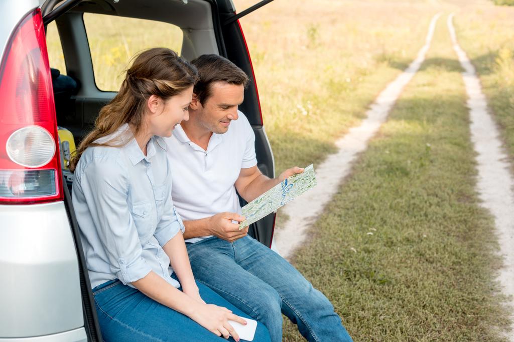 усміхнена доросла пара сидить у багажнику автомобіля і дивиться на карту під час поїздки на машині
 - Фото, зображення