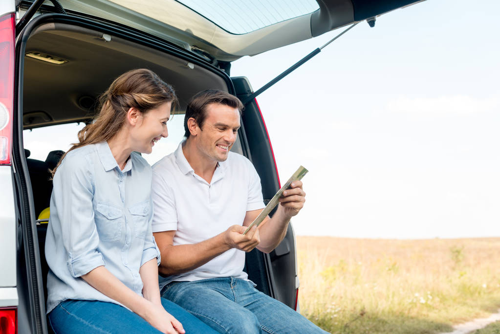 szczęśliwa para dorosłych siedzi w bagażniku samochodu i patrząc na mapę mając podróż samochodem - Zdjęcie, obraz