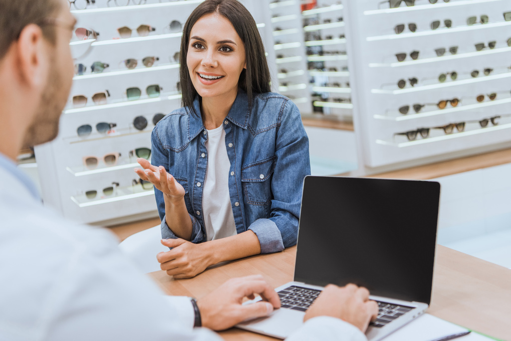 gedeeltelijke weergave van mannelijke optometrist bezig met laptop terwijl woman wijzend met de hand in ophthalmic winkel - Foto, afbeelding