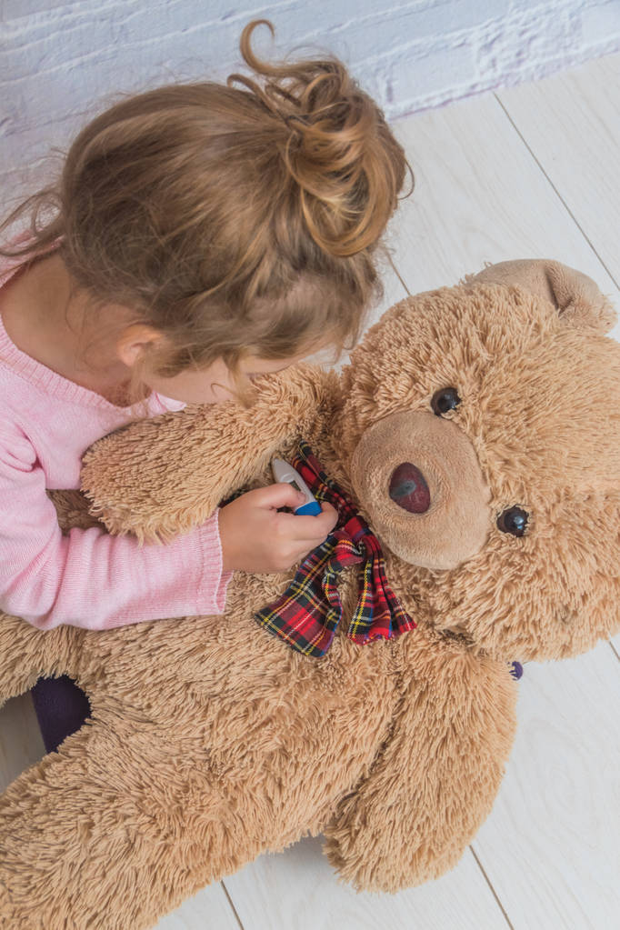 Девочка, ребенок измеряет температуру игрушечного медведя, прислонившись к стене из белых брикетов
 - Фото, изображение