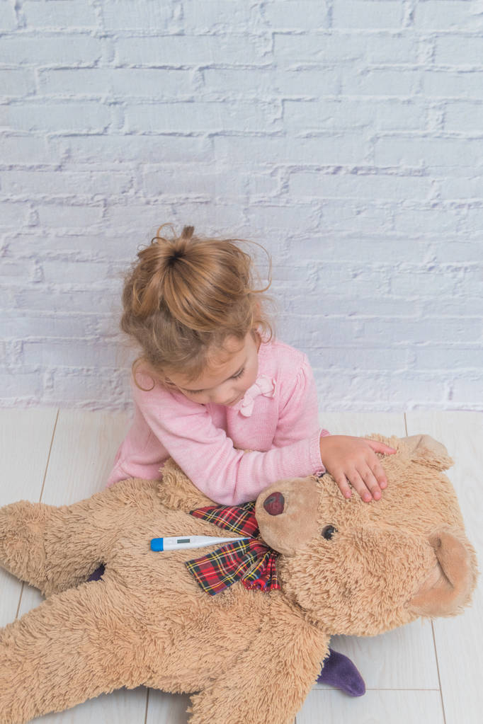 la niña, el niño mide la temperatura de un oso de juguete, contra una pared de ladrillo blanco
 - Foto, Imagen
