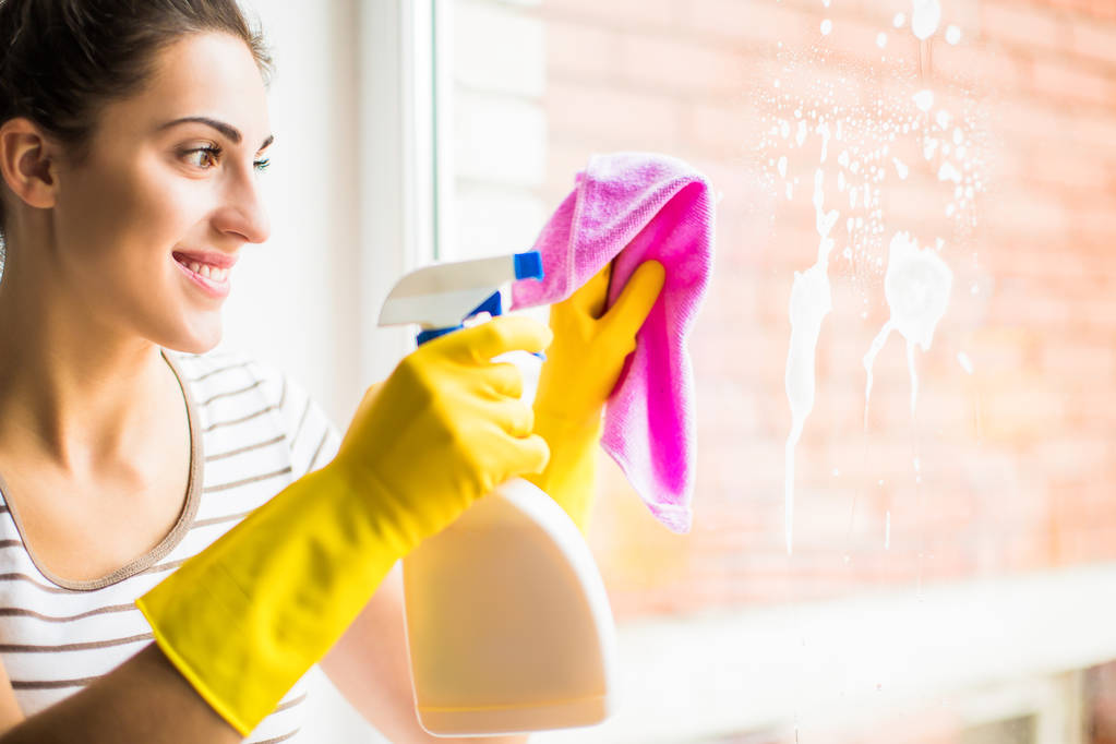 De vrolijke jongedame in handschoenen wassen het venster thuis - Foto, afbeelding
