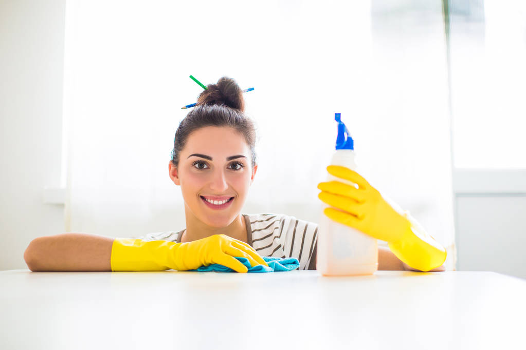 Retrato de la joven sonriente con guantes amarillos que sostiene el trapo y el detergente y se sienta cerca de la mesa en la habitación
 - Foto, Imagen