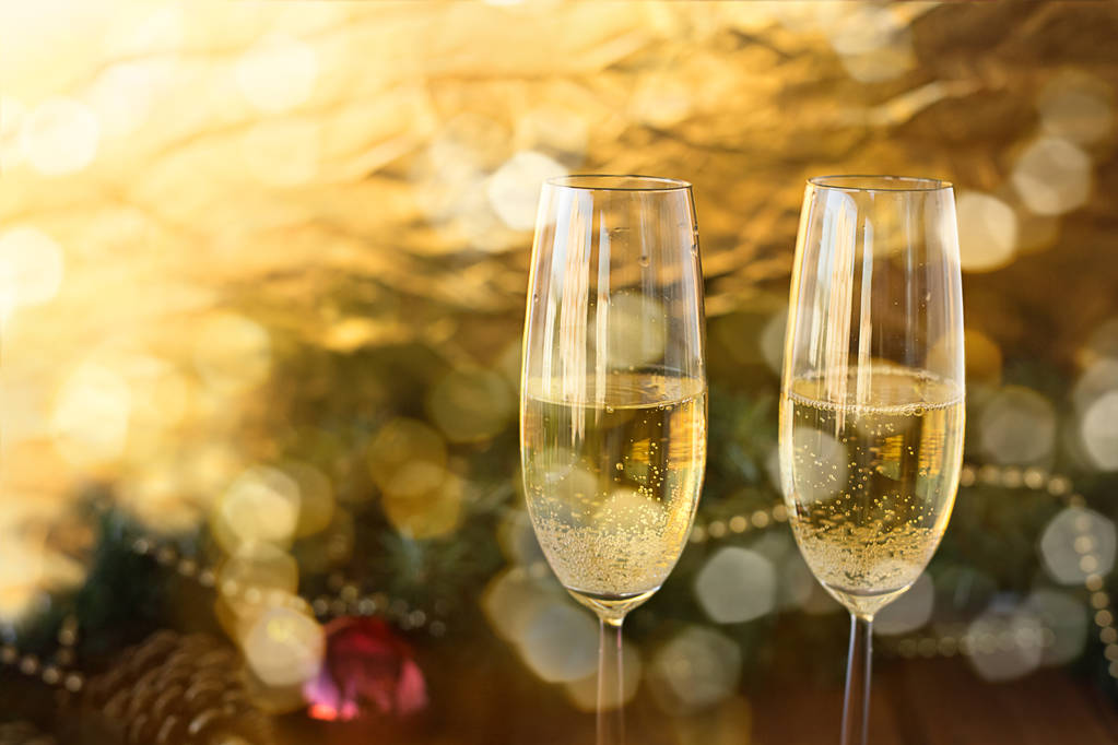 два новогодних бокала с шампанским на деревянном столе и золотом на белом фоне и ветки украшенной елки в сиянии с голубым
 - Фото, изображение