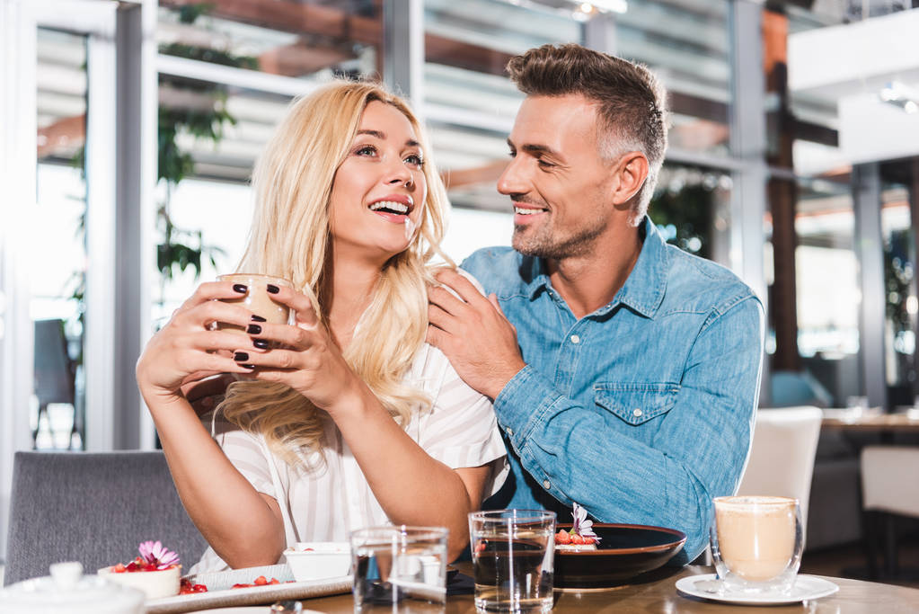 хлопець обіймає подругу сміється і вона тримає чашку кави за столом в кафе
 - Фото, зображення