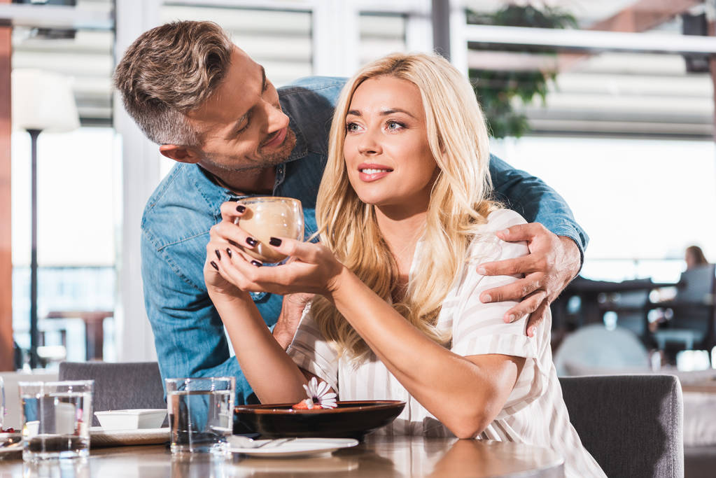 handsome boyfriend hugging girlfriend near table in cafe, she holding glass of coffee - Photo, Image