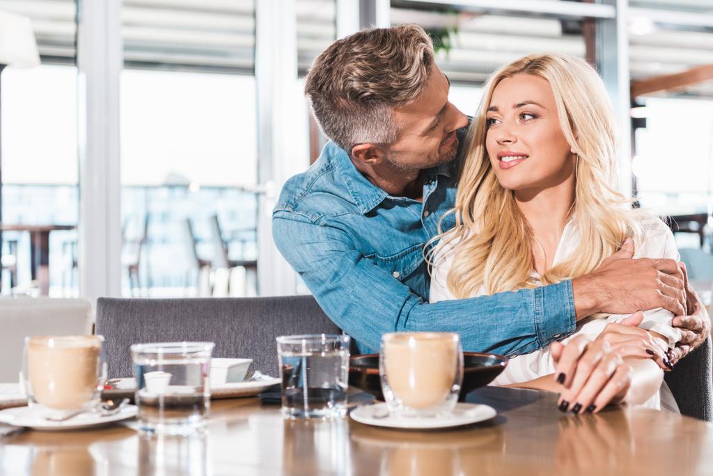knappe vriendje knuffelen aantrekkelijke vriendin aan tafel in café - Foto, afbeelding