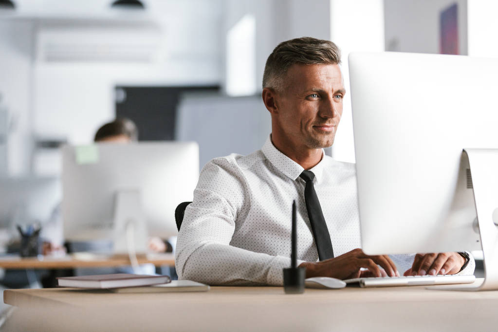 Imagem do empregado bem sucedido 30 anos vestindo camisa branca e gravata sentado na mesa no escritório e trabalhando no computador
 - Foto, Imagem