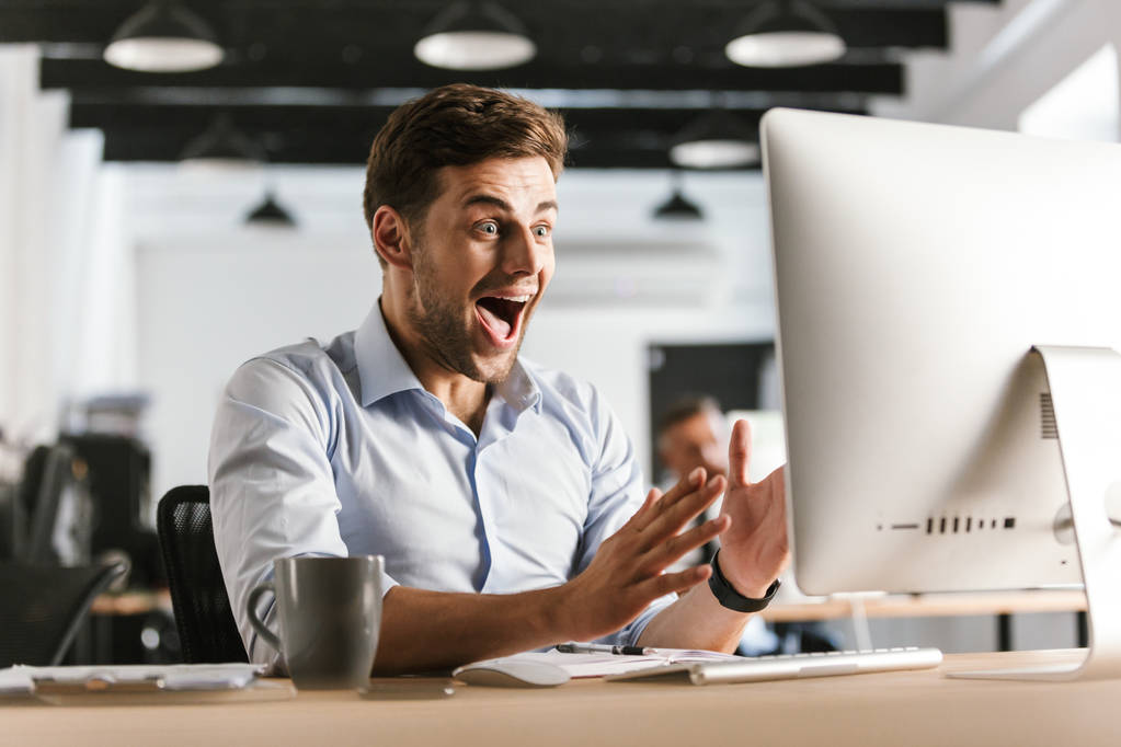 Homem de negócios gritando chocado usando o computador enquanto sentado à mesa no escritório
 - Foto, Imagem