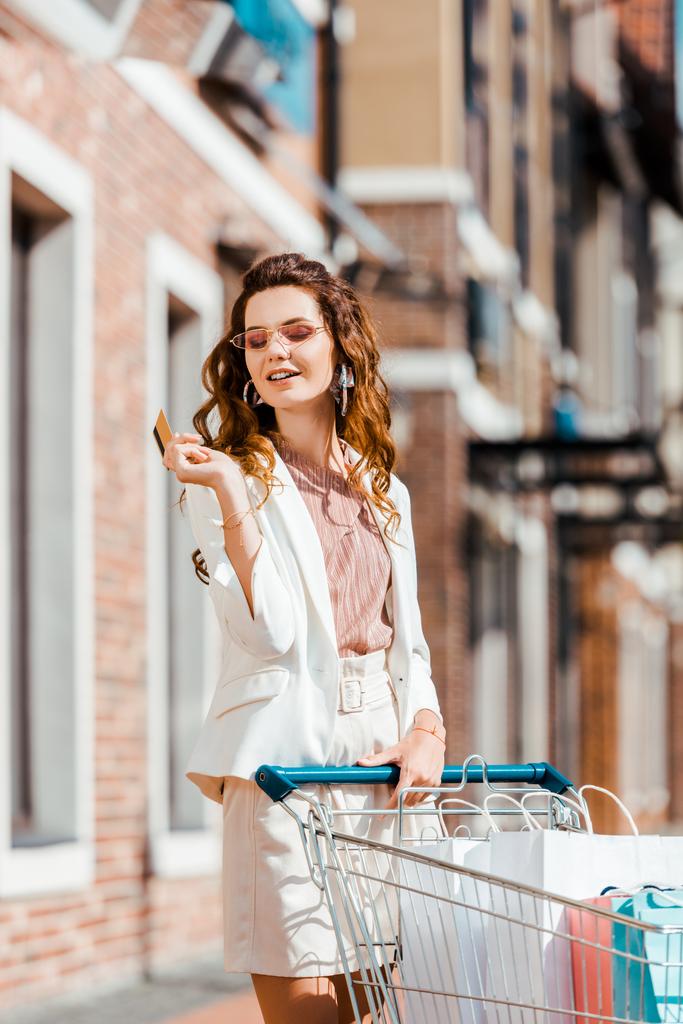 glückliche junge Frau mit Einkaufswagen voller Papiertüten mit Kreditkarte - Foto, Bild
