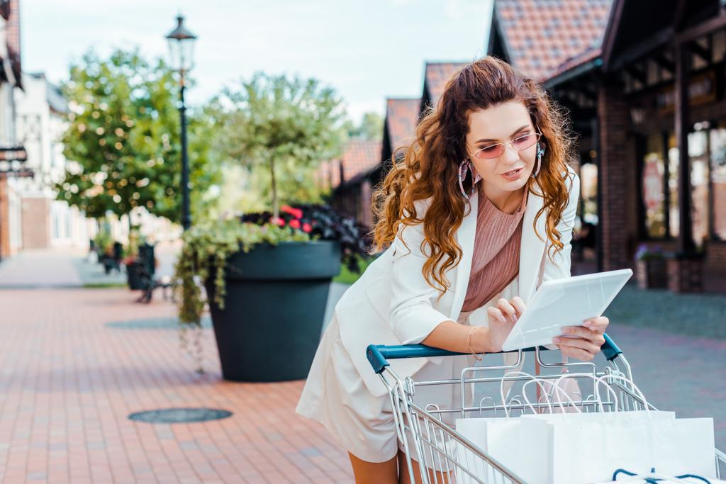 smiling stylish woman with shopping cart full of paper bags using tablet - Photo, Image