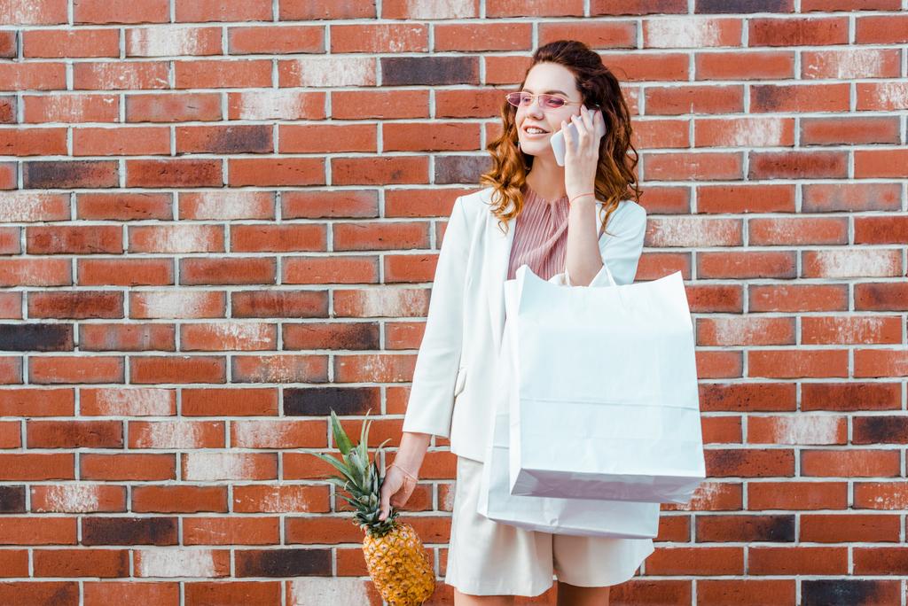 bela jovem com abacaxi e sacos de compras falando por telefone na frente da parede de tijolo
 - Foto, Imagem