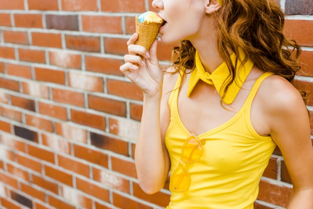 Schnappschuss einer jungen Frau in gelber Kleidung, die vor einer Ziegelwand Eis isst - Foto, Bild