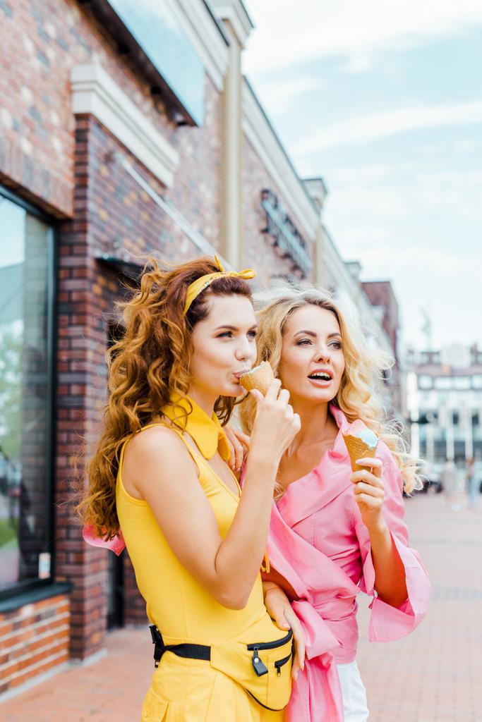 όμορφες νεαρές γυναίκες με πολύχρωμα ρούχα τρώγοντας παγωτό σε κώνους βαφλών στην οδό - Φωτογραφία, εικόνα