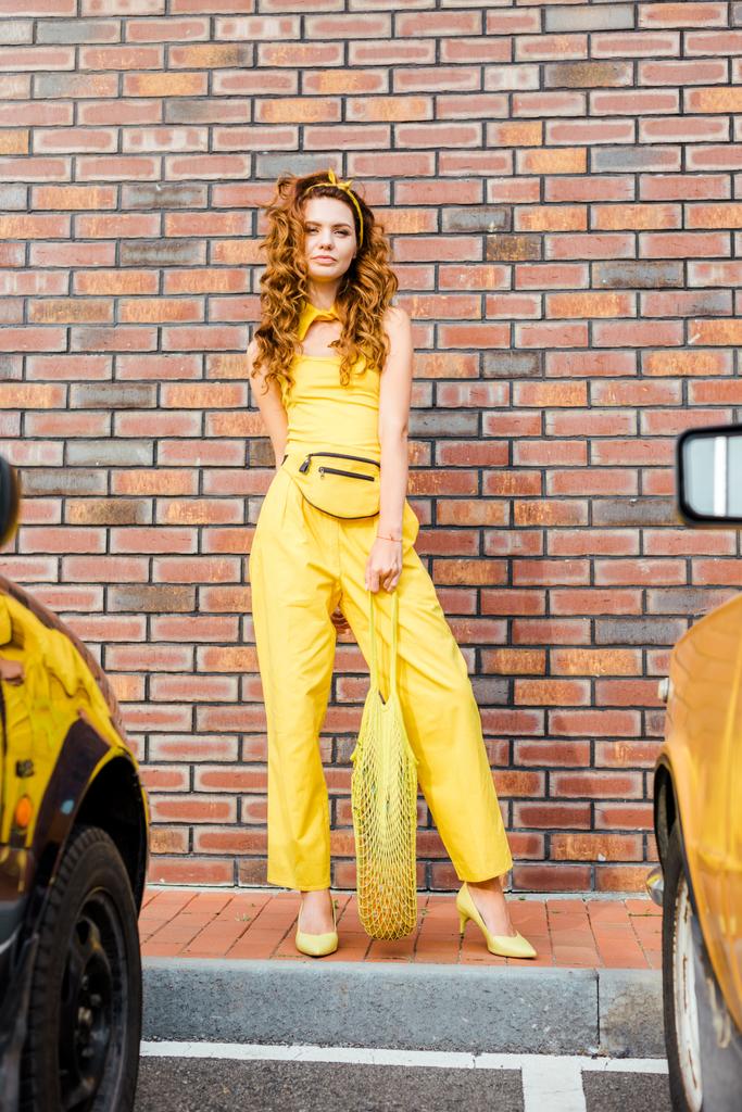 hermosa mujer joven en ropa amarilla con piña en bolsa de hilo de pie en el aparcamiento delante de la pared de ladrillo
 - Foto, imagen