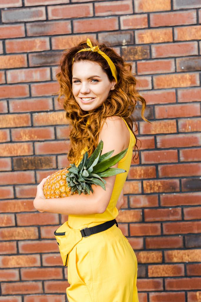Gelukkig jonge vrouw in gele kleding ananas voor bakstenen muur te houden en te kijken naar camera - Foto, afbeelding