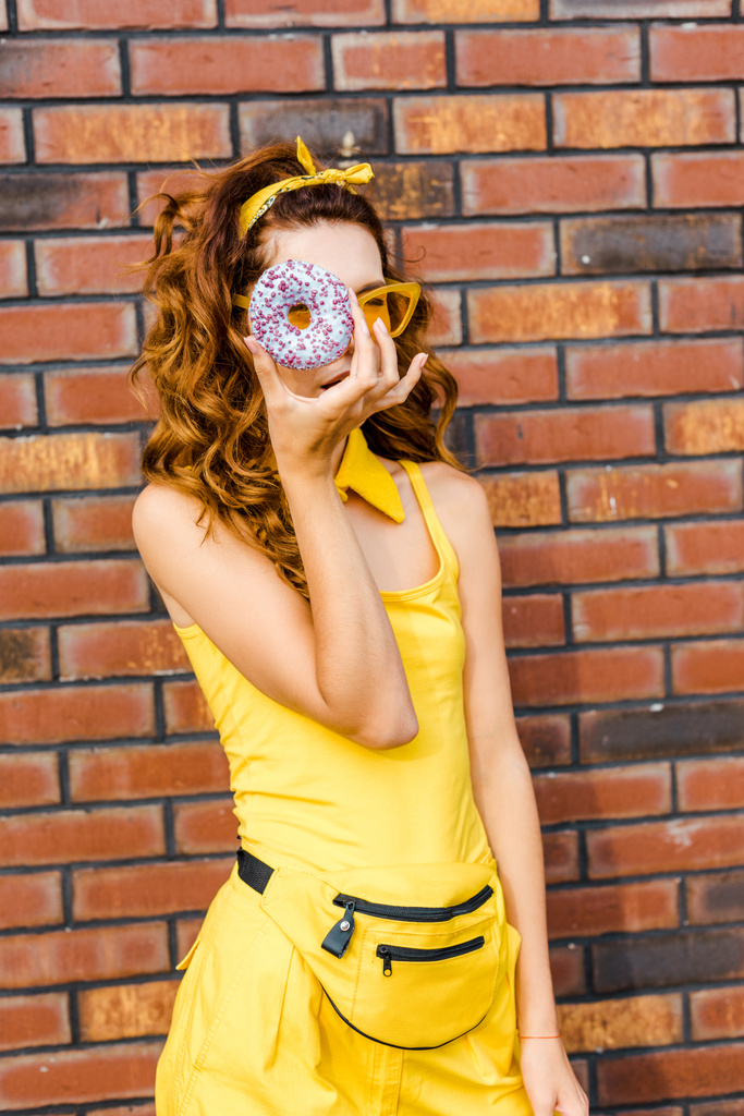 attraktive junge Frau in gelben Kleidern blickt durch Donut vor Ziegelwand in die Kamera - Foto, Bild