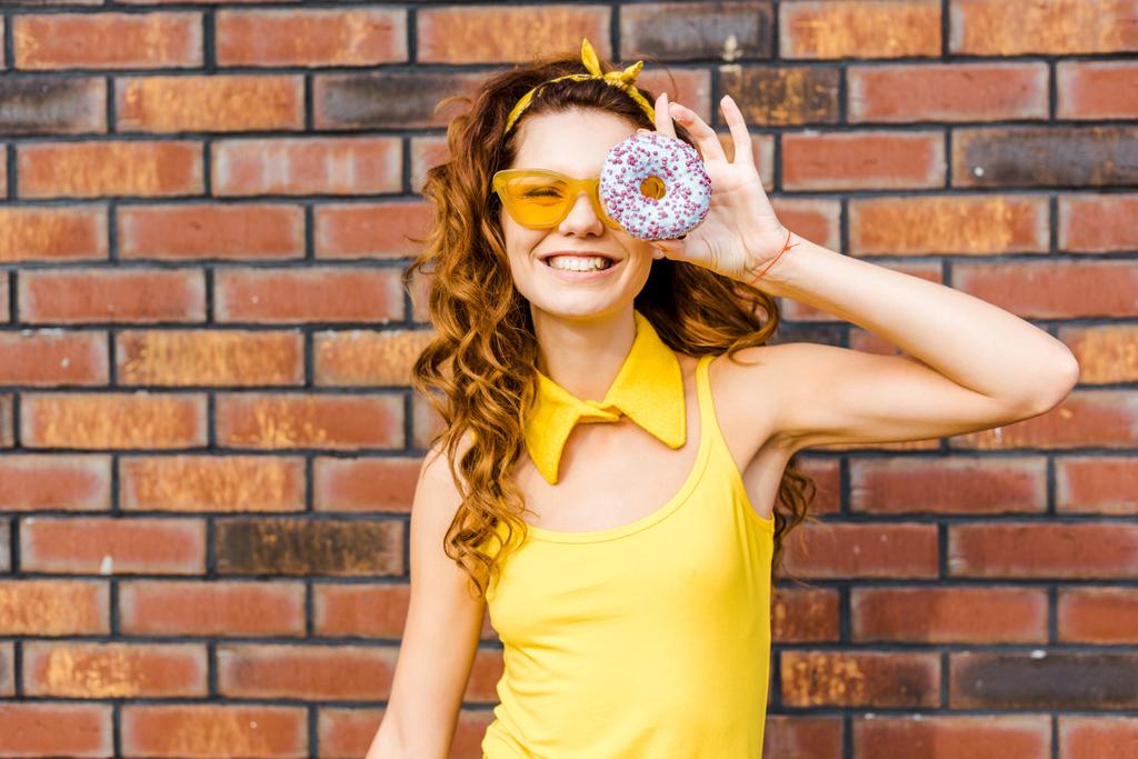 smiling young woman in yellow clothes looking at camera through donut in front of brick wall - Photo, Image