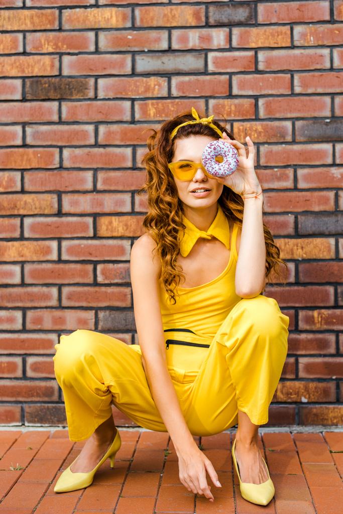 όμορφη νεαρή γυναίκα με κίτρινα ρούχα βλέπουν φωτογραφική μηχανή μέσω ντόνατ που κάθεται μπροστά από τον τοίχο από τούβλα - Φωτογραφία, εικόνα