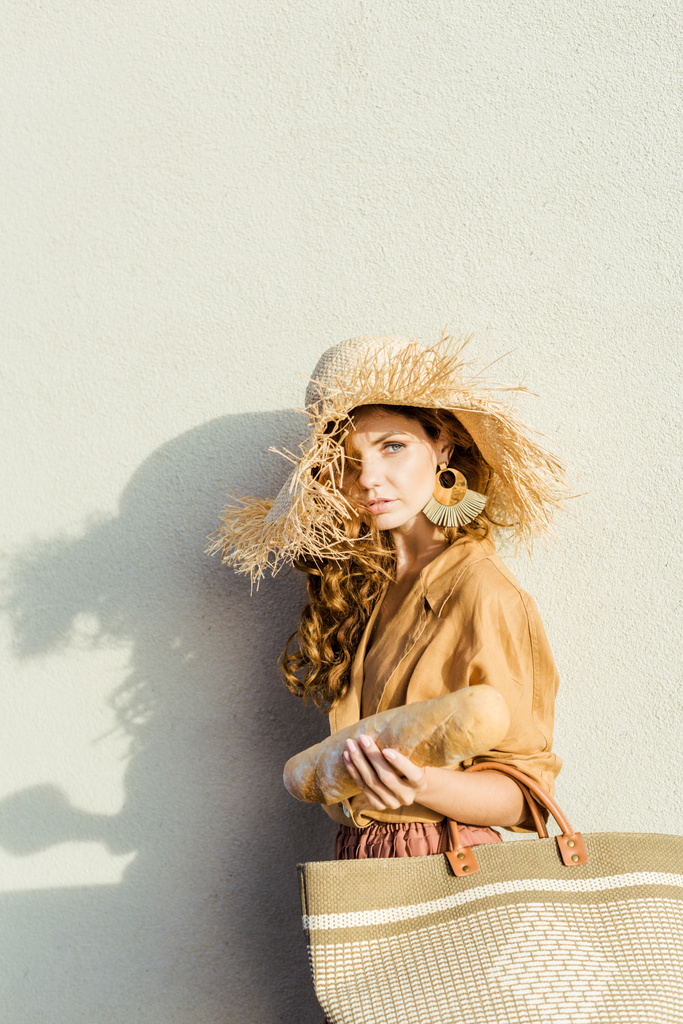 giovane donna alla moda in cappello di paglia con baguette in piedi davanti al muro bianco
 - Foto, immagini