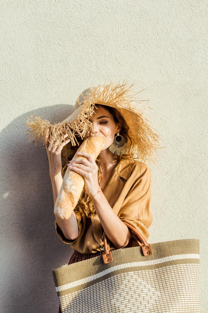 hasır şapka yeme bir baget beyaz duvarın önünde duran güzel genç kadın - Fotoğraf, Görsel