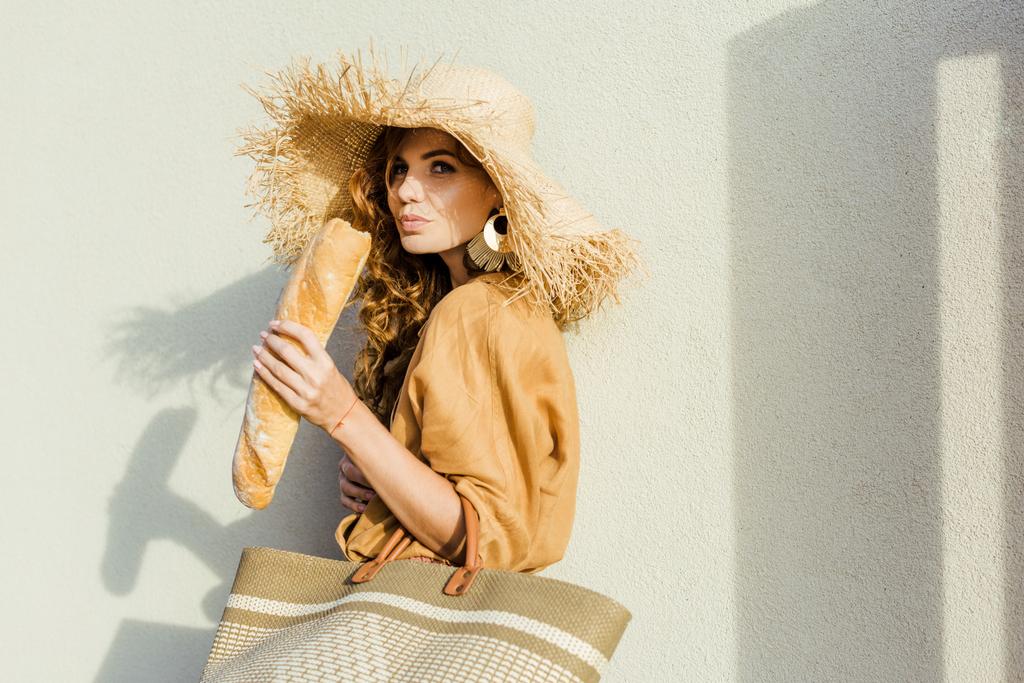 stylish young woman in straw hat standing in front of white wall and eating french baguette  - Photo, Image
