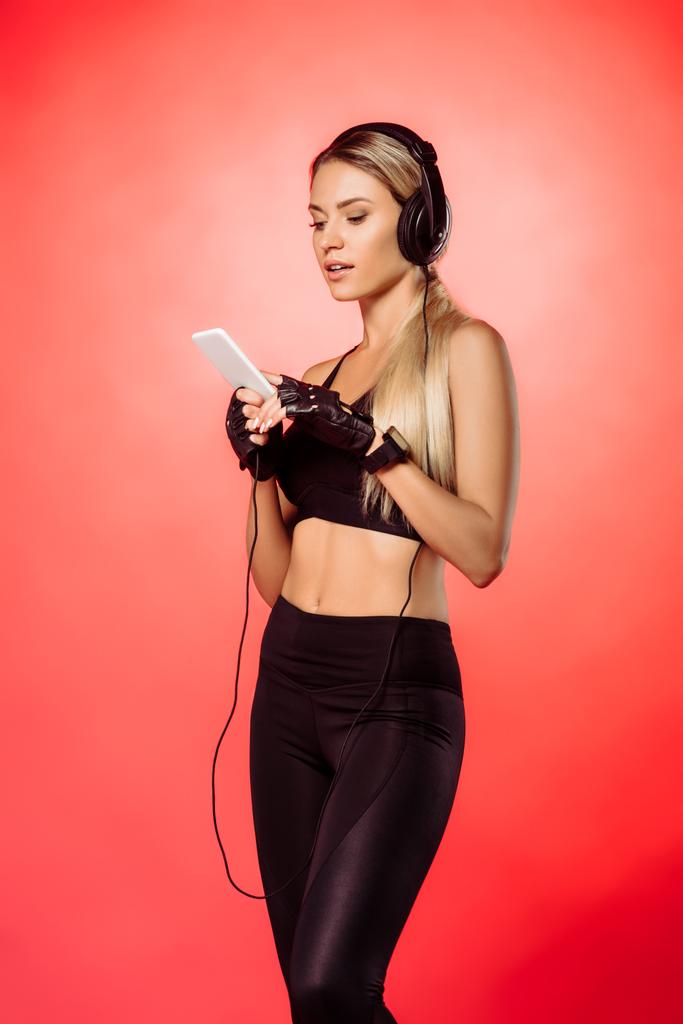 привлекательная спортсменка слушает музыку со смартфоном, изолированным на красном
 - Фото, изображение