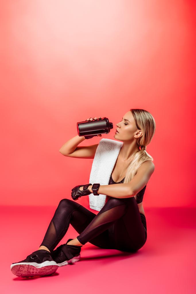 привлекательная спортсменка сидит с полотенцем и пьет белковые добавки из спортивной бутылки на красном
 - Фото, изображение