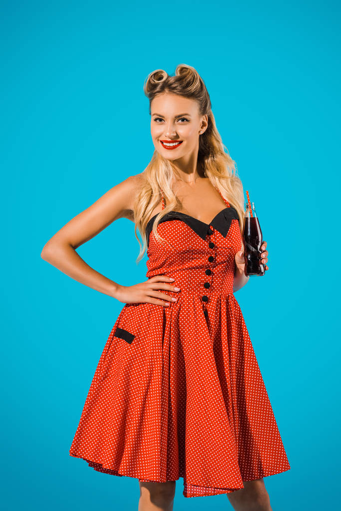 portrait of smiling pin up woman in vintage dress with soda drink in glass bottle on blue background - Photo, Image