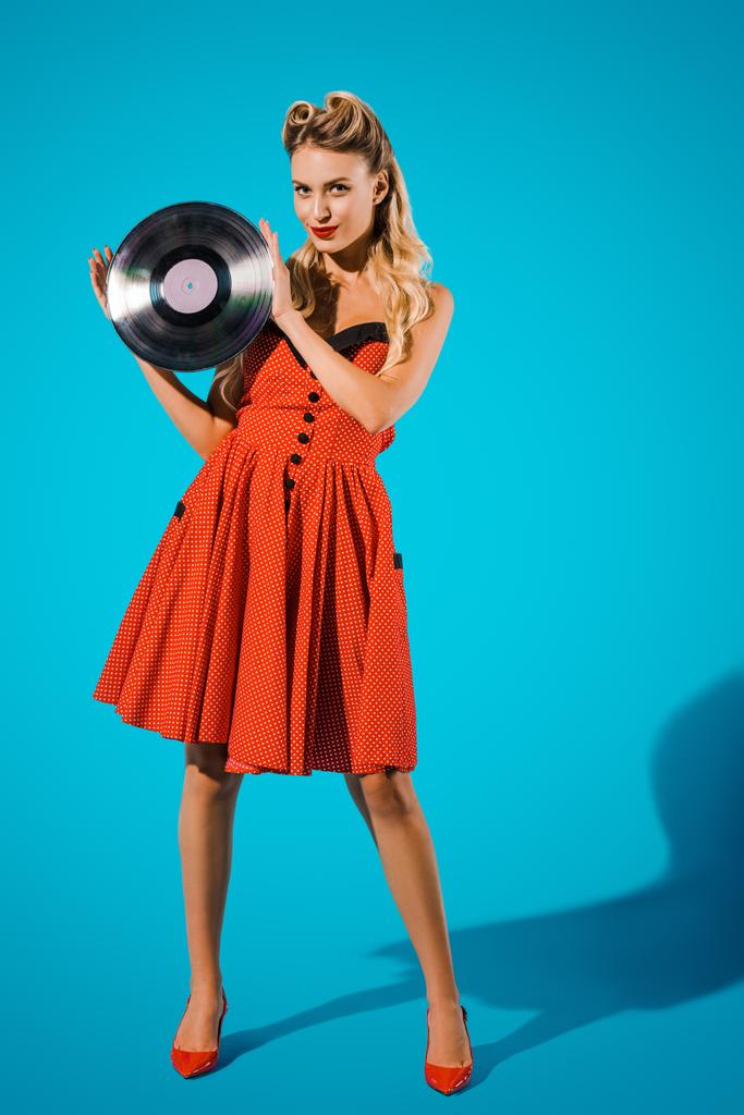young pin up woman in stylish vintage dress with vinyl record on blue backdrop - Photo, Image