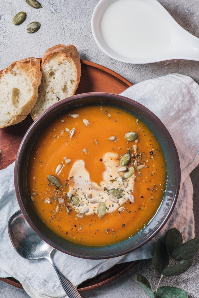 verhoogde weergave van pompoen crème soep met zaden in een kom en brood op houten bord op tafel  - Foto, afbeelding