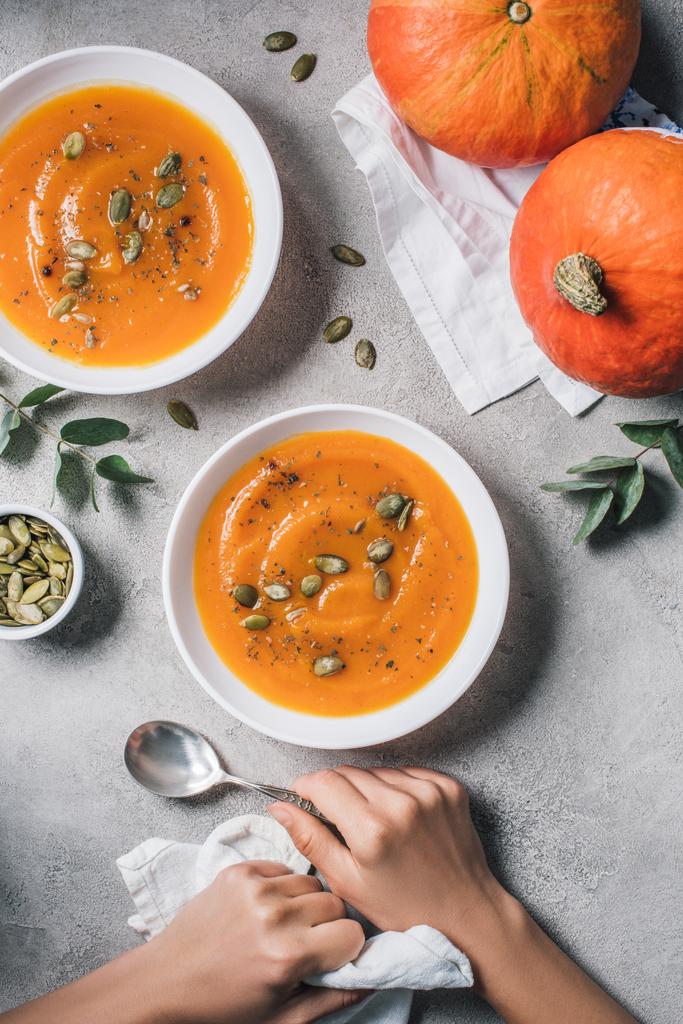 partial view of woman sitting at table with pumpkins and plates with pumpkin soup and seeds  - Photo, Image