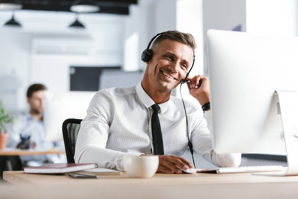 Foto dell'operatore sorridente uomo 30 anni che indossa abiti da ufficio e auricolare seduto al computer in call center
 - Foto, immagini