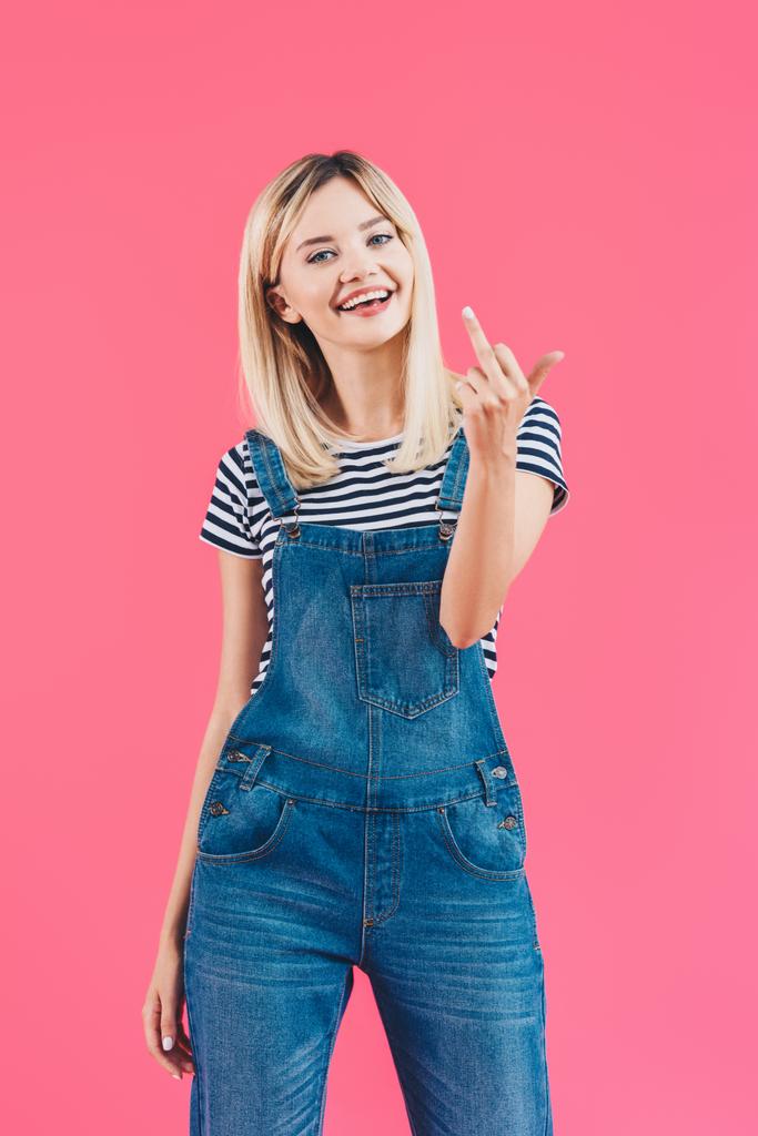 smiling beautiful girl in denim overall showing middle finger isolated on pink - Photo, Image