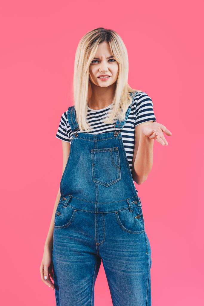 grimacing girl in denim overall gesturing and looking at camera isolated on pink - Photo, Image